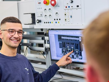 junger-mann-zeigt-auf-monitor junger Mann erklärte einem anderen Auszubildenden etwas am Monitor