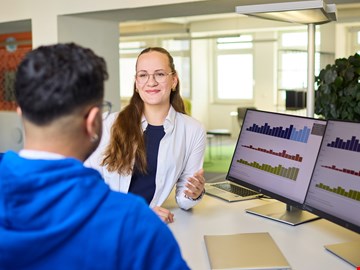 mann-frau-grafiken Mann und Frau im Gespräch am Arbeitsplatz vor zwei Monitoren mit Grafiken